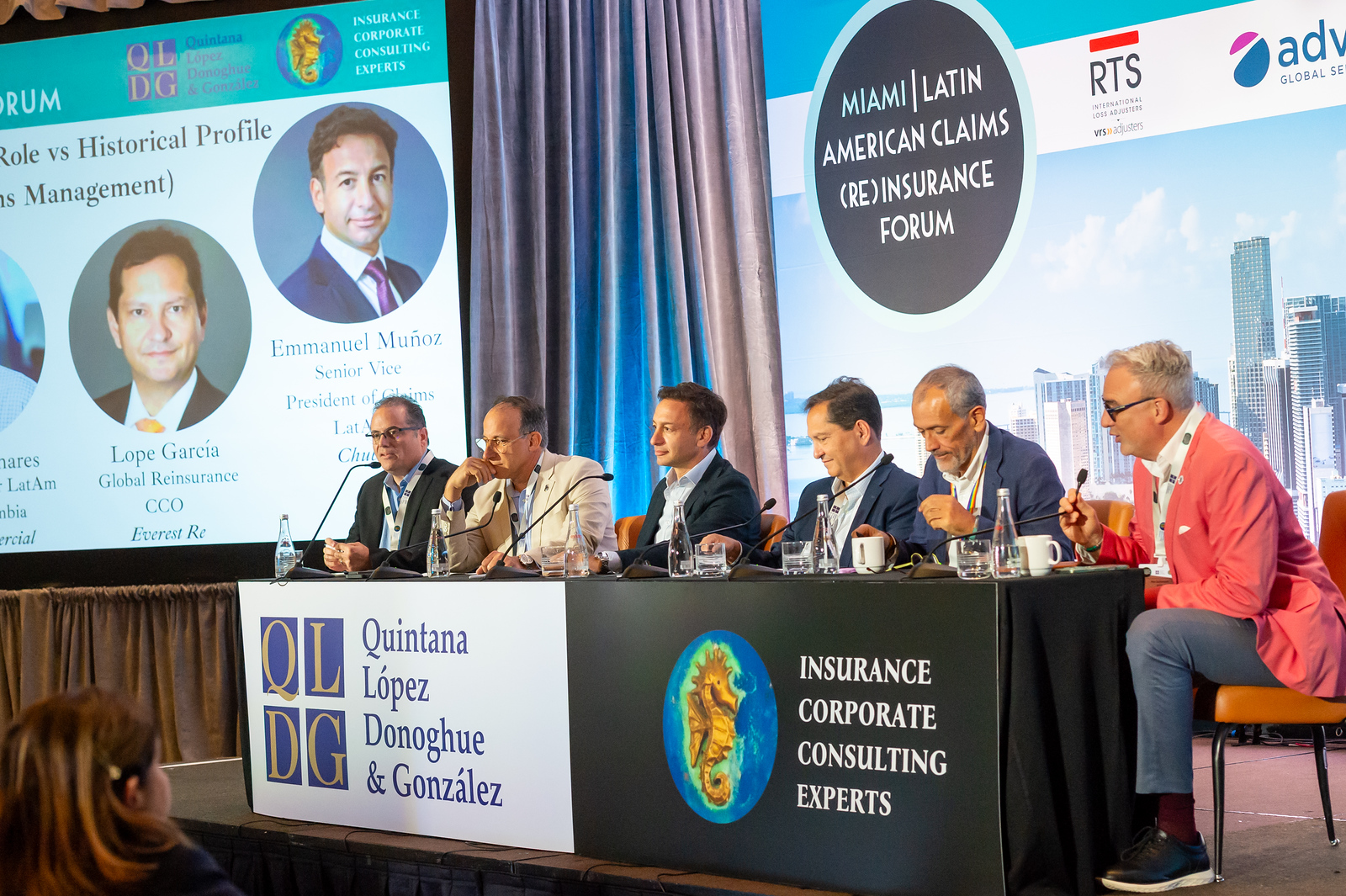 Six male panelists seated at a long table with microphones and water bottles, participating in the Miami Latin American Claims (Re)Insurance Forum, with a backdrop showing portraits of Lope GarcĂa and Emmanuel Muñoz and logos for Quintana LĂłpez Donoghue & González and corporate consulting experts.